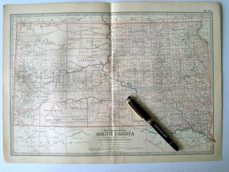 Może przedstawiać: Antyczna mapa Dakoty Południowej, pokazująca granice hrabstw i miasta. Mapa jest otwarta na stronie w oprawionym atlasie, z czarnym długopisem leżącym na stronie.