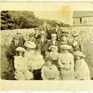 May include: A vintage black and white photograph of a group of people, mostly women, wearing hats, gathered outdoors. The group is standing in front of a stone wall with a building in the background.