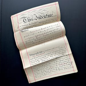 May include: An antique document, titled "This Indenture," is open and displayed against a dark background. The document features ornate calligraphy and pink border lines. The text is in black ink.