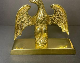 May include: A gold-colored metal eagle sculpture with outstretched wings. The eagle is perched on a rectangular base. The sculpture has detailed feather and wing patterns. The background is a dark gray.