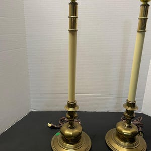 May include: Two vintage brass table lamps with off-white columns and ornate bases. Each lamp has a gold-colored bulb socket and a brown electrical cord. A measuring tape is visible at the bottom.