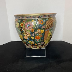 Puede incluir: Un macetero grande, redondo, de porcelana negra y blanca con un diseño floral. El macetero tiene un borde dorado y está decorado con una variedad de flores, incluyendo rosas, peonías y crisantemos. El macetero está sentado en un soporte de acrílico transparente.