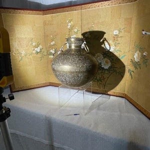 Puede incluir: Un jarrón grande, adornado, de metal de color dorado con intrincados diseños florales. El jarrón está sobre un soporte de acrílico transparente.