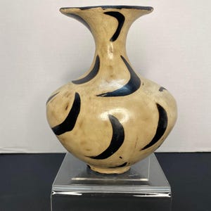 May include: A beige ceramic vase with a flared neck and rounded body. The vase is adorned with black, crescent-shaped designs. The vase is displayed on a clear acrylic stand against a white background.
