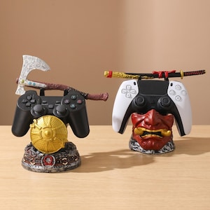 Pode incluir: Dois suportes decorativos para comandos de jogos. Um apresenta um comando preto com um escudo dourado e um machado, o outro um comando branco com uma máscara vermelha e uma katana. Ambos estão numa superfície de madeira.