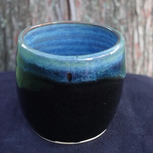 May include: A small, round ceramic cup with a black base and a blue and green glaze. The cup is glazed on the inside and outside.