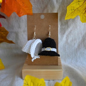 May include: A pair of handmade crochet earrings. One earring is a white bonnet, the other is a black pilgrim hat with a white and yellow band. The earrings are displayed on a brown card and wooden stand, with fall foliage in the background.