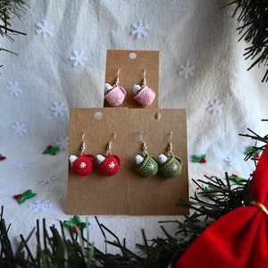 May include: Crocheted Christmas-themed earrings in pink, red, and green, shaped like mugs. Each pair is displayed on a brown card. The earrings have white accents and a small red star on the red mugs. The background includes snowflakes and a red bow.