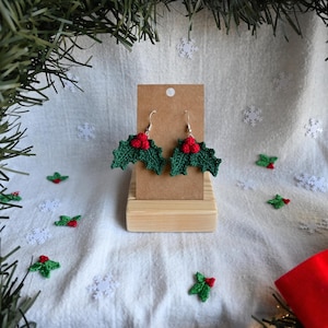 May include: Handmade green crochet holly leaf earrings with red berry details. The earrings are presented on a small wooden stand against a white backdrop, decorated with snowflakes and holly leaves, creating a festive Christmas theme.
