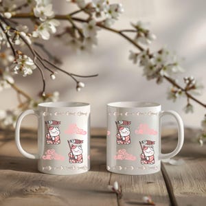 Könnte beinhalten: Zwei weiße Keramikbecher mit einem sich wiederholenden Muster einer Cartoon-Katze, die eine Fischflagge, rosa Wolken und Fischgräten hält. Die Becher stehen auf einer Holzoberfläche, mit weißen Blumen im Hintergrund.