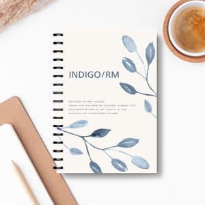 Può includere: Un taccuino a spirale color crema con il testo "INDIGO/RM" e illustrazioni ad acquerello di foglie blu. Il testo del taccuino recita: "RECORD OF RM INDIGO FROM THE COLORS OF NATURE, HUMAN, ETC. DOCUMENTATION OF MY YOUTH IN THE MOMENT OF INDEPENDENT PHASE."