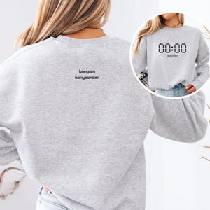 Könnte beinhalten: Ein hellgraues Sweatshirt mit dem Text "00:00" in einer Digitaluhr-Schriftart auf der Vorderseite. Auf der Rückseite des Sweatshirts steht der Text "bangran sonyeondan". Das Sweatshirt ist ein lässiger Stil.