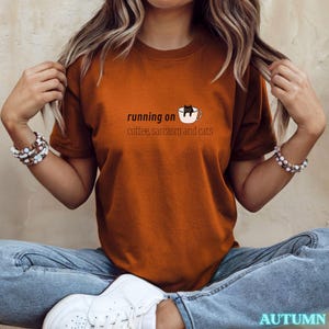 Könnte beinhalten: Ein rostfarbenes T-Shirt mit dem Text "running on coffee, sarcasm and cats" und einer Grafik einer Katze in einer Kaffeetasse. Das Wort "AUTUMN" steht unten rechts.