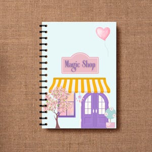 Könnte beinhalten: Ein spiralgebundenes Notizbuch mit hellblauem Einband. Die Illustration zeigt ein Zaubergeschäft mit einer gelb-weiß gestreiften Markise, lila Türen und einem rosa Schild mit der Aufschrift "Magic Shop". Ein rosa herzförmiger Ballon schwebt darüber.