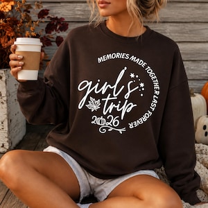 Puede incluir: Una sudadera de cuello redondo en color marrón oscuro con texto impreso en blanco que dice "MEMORIES MADE TOGETHER LAST FOREVER" en arco, junto a "girl's trip" en letra cursiva, una hoja de arce, una pequeña calabaza y el año "2026".