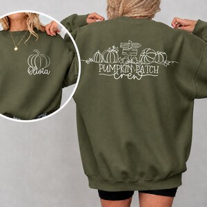 May include: An olive green crewneck sweatshirt featuring a white line art pumpkin and the name Olivia on the front left chest. The back displays a white graphic of pumpkins, a sign reading Hand Picked, and the text Pumpkin Patch crew.