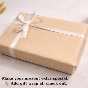 May include: A rectangular gift box wrapped in tan paper and tied with a white satin ribbon. The box is on a white surface, with a small vase of dried flowers, a candle, a pen, and a small dish with jewelry.