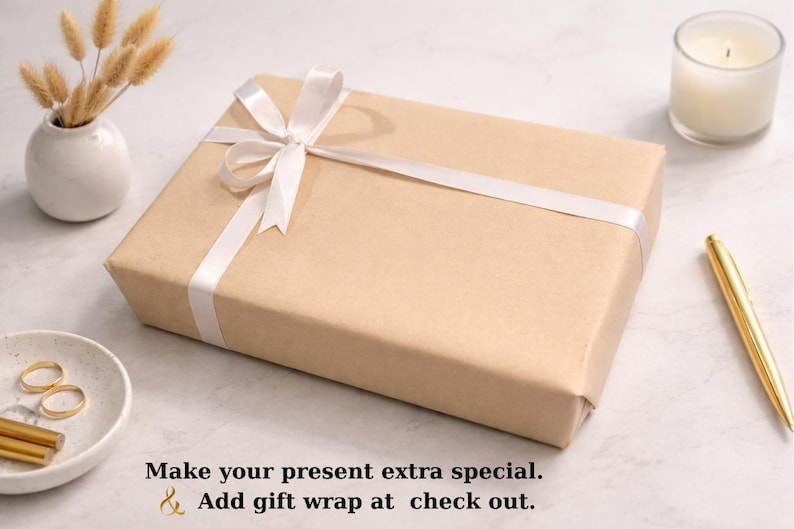 May include: A rectangular gift box wrapped in tan paper and tied with a white satin ribbon. The box is on a white surface, with a small vase of dried flowers, a candle, a pen, and a small dish with jewelry.
