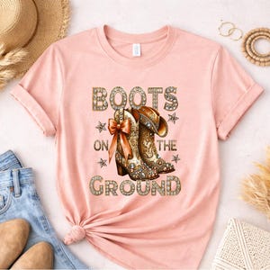 May include: A light pink t-shirt featuring the text "BOOTS ON THE GROUND" in a decorative font. The graphic includes a pair of detailed cowboy boots with a bow and stars. The shirt is folded, with a straw hat and jeans visible.