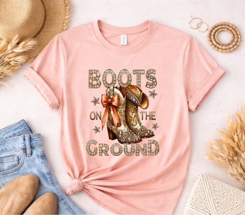 May include: A light pink t-shirt featuring the text "BOOTS ON THE GROUND" in a decorative font. The graphic includes a pair of detailed cowboy boots with a bow and stars. The shirt is folded, with a straw hat and jeans visible.