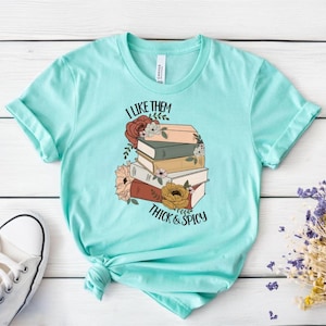 Op de afbeelding: Een turquoise t-shirt met een afbeelding van een stapel boeken met bloemen en de tekst "I like them thick & spicy".