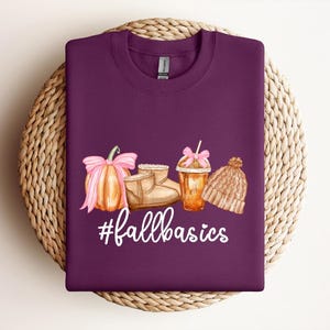 May include: A dark purple sweatshirt with a watercolor design of fall essentials: a pumpkin with a pink bow, boots, a coffee drink, and a knit hat. The text "#fallbasics" is written in white.