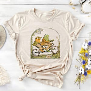 以下が含まれることがあります： ベージュのTシャツに、自転車に乗った2匹のカエルのグラフィックがプリントされています。カエルは緑と茶色で、服を着ています。自転車は赤で、前にバスケットが付いています。