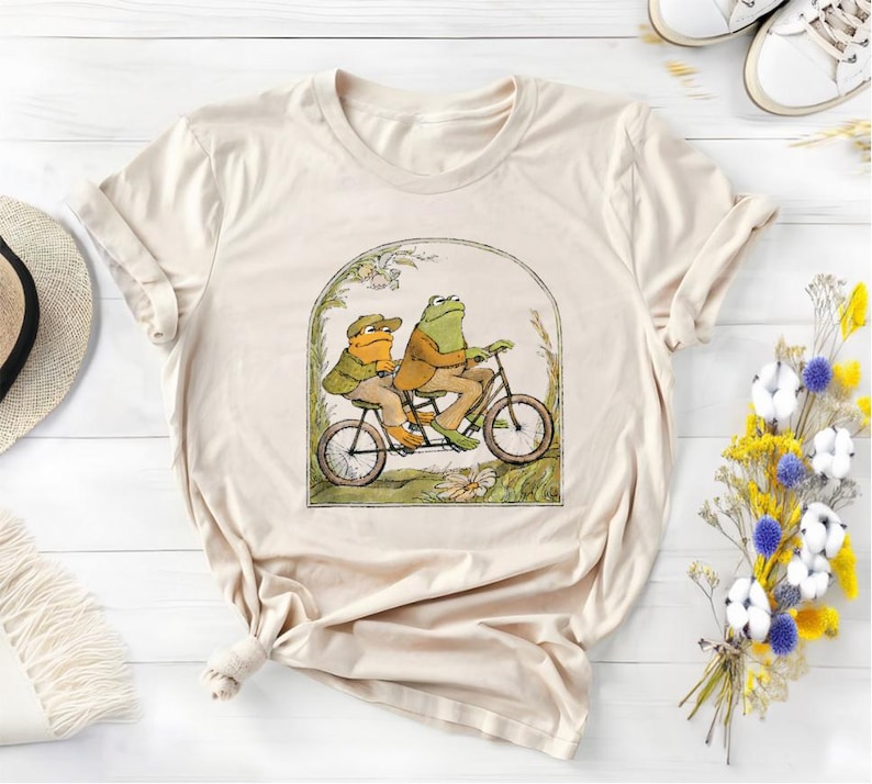 以下が含まれることがあります： ベージュのTシャツに、自転車に乗った2匹のカエルのグラフィックがプリントされています。カエルは緑と茶色で、服を着ています。自転車は赤で、前にバスケットが付いています。