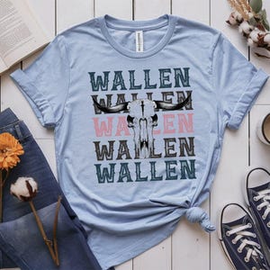 K&ouml;nnte beinhalten: Hellblaues T-Shirt mit dem Wort "WALLEN" vertikal gestapelt, mit einer Bullensch&auml;del-Grafik. Das Wort "WALLEN" ist in Schwarz, Rosa und T&uuml;rkis gehalten. Das Shirt wird mit blauen Jeans und marineblauen Sneakers gestylt.