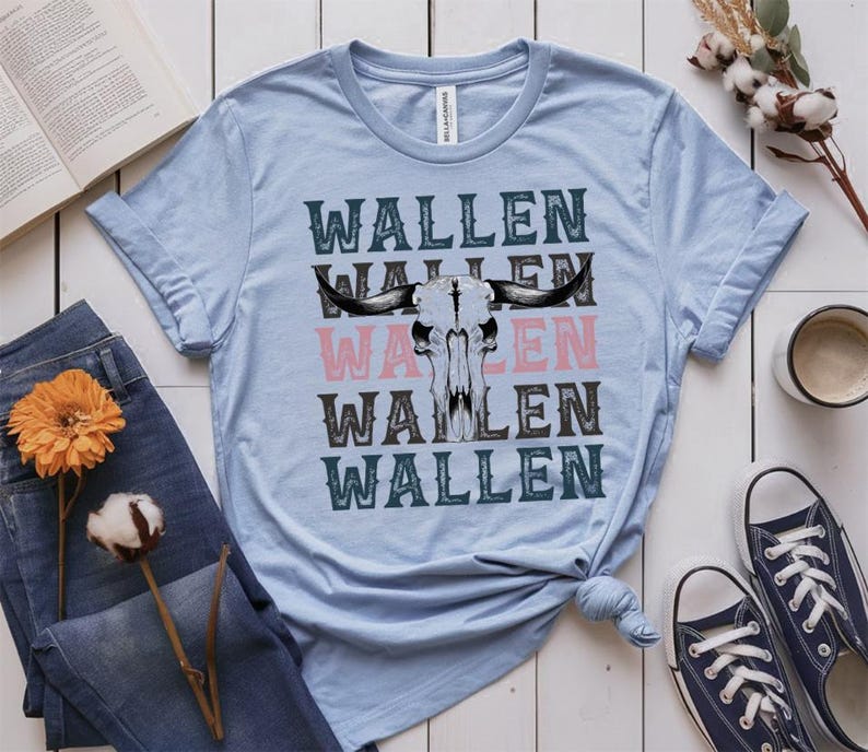 K&ouml;nnte beinhalten: Hellblaues T-Shirt mit dem Wort "WALLEN" vertikal gestapelt, mit einer Bullensch&auml;del-Grafik. Das Wort "WALLEN" ist in Schwarz, Rosa und T&uuml;rkis gehalten. Das Shirt wird mit blauen Jeans und marineblauen Sneakers gestylt.