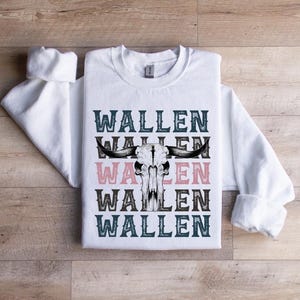 K&ouml;nnte beinhalten: Wei&szlig;es Sweatshirt mit dem Wort "WALLEN" in T&uuml;rkis und Rosa, mit einem Bullensch&auml;del-Motiv. Das Sweatshirt liegt flach auf einer Holzoberfl&auml;che.