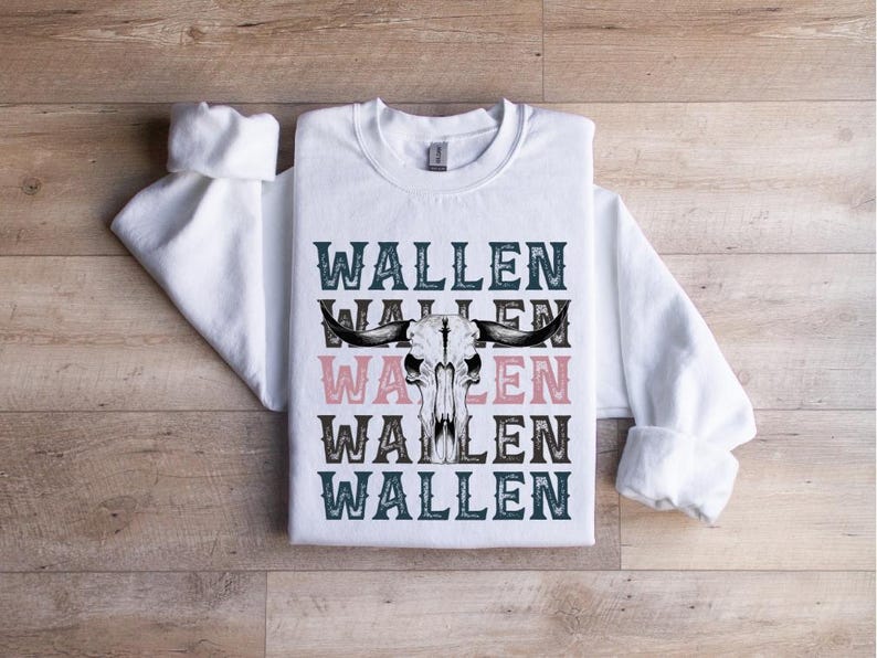 K&ouml;nnte beinhalten: Wei&szlig;es Sweatshirt mit dem Wort "WALLEN" in T&uuml;rkis und Rosa, mit einem Bullensch&auml;del-Motiv. Das Sweatshirt liegt flach auf einer Holzoberfl&auml;che.