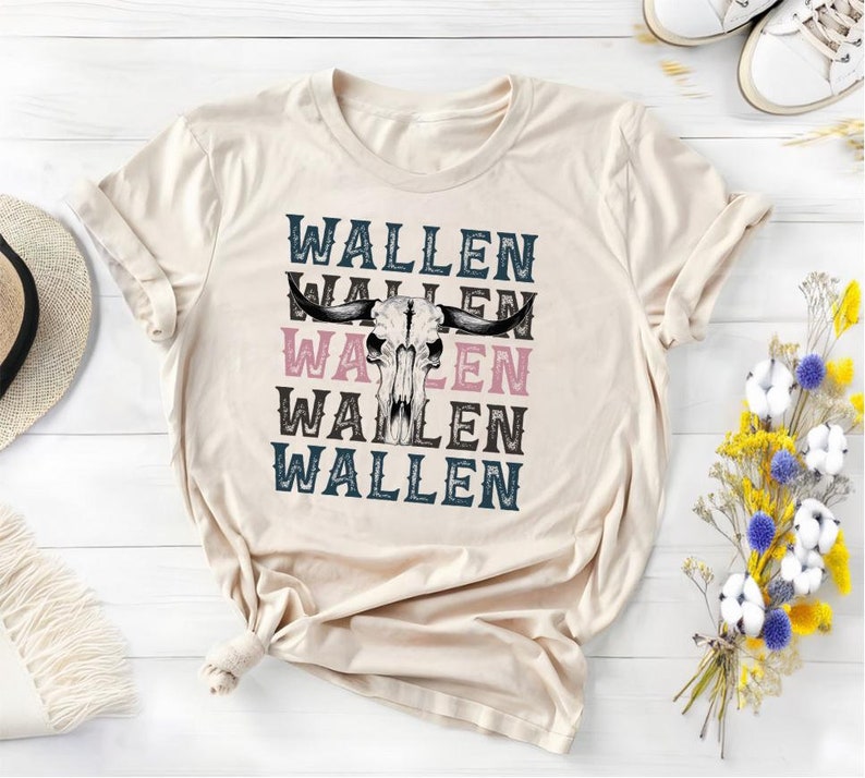 K&ouml;nnte beinhalten: Cremefarbenes T-Shirt mit dem Wort "WALLEN" viermal &uuml;bereinander und einer Bullensch&auml;del-Grafik. Der Text wechselt zwischen Blau, Rosa und Braun. Das Shirt liegt gefaltet auf einer wei&szlig;en Holzoberfl&auml;che.