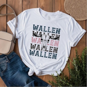 K&ouml;nnte beinhalten: Wei&szlig;es T-Shirt mit einem Bullensch&auml;del-Motiv und dem Namen "WALLEN" in Schwarz, Rosa und T&uuml;rkis &uuml;bereinander angeordnet. Das Shirt wird mit einer blauen Jeans und einer beigen Handtasche kombiniert.