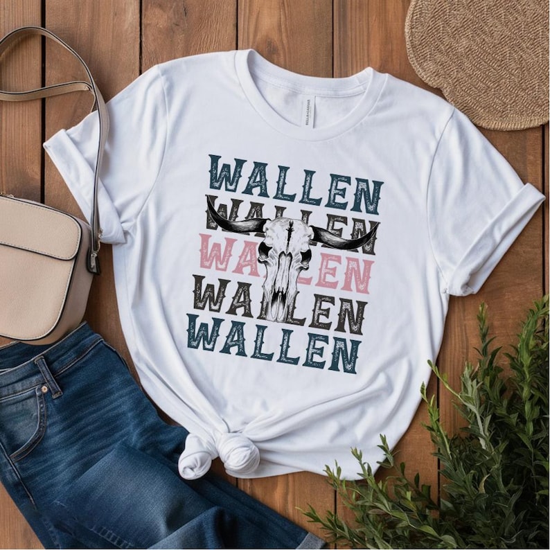 K&ouml;nnte beinhalten: Wei&szlig;es T-Shirt mit einem Bullensch&auml;del-Motiv und dem Namen "WALLEN" in Schwarz, Rosa und T&uuml;rkis &uuml;bereinander angeordnet. Das Shirt wird mit einer blauen Jeans und einer beigen Handtasche kombiniert.