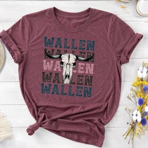 K&ouml;nnte beinhalten: Burgunderfarbenes T-Shirt mit umgekrempelten &Auml;rmeln. Es zeigt eine Grafik eines Bullensch&auml;dels und den Namen "WALLEN" in Blau, Rosa und Schwarz &uuml;bereinander. Das T-Shirt ist aus weichem Material. Das Design ist ein Statement.