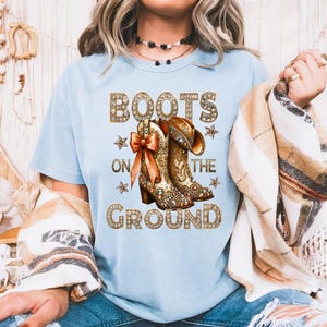 May include: Light blue t-shirt with the text "BOOTS ON THE GROUND" in a sparkling gold font. The graphic features a pair of detailed cowboy boots with a bow. The shirt is a casual wear item.