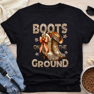 May include: Black t-shirt featuring a graphic of embellished cowboy boots with the text "BOOTS ON THE GROUND" in a rhinestone-like font. The boots are brown and gold with a decorative bow.