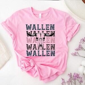 K&ouml;nnte beinhalten: Ein pinkfarbenes T-Shirt mit dem Wort "WALLEN" viermal wiederholt und einer Bullensch&auml;del-Grafik in der Mitte. Der Text ist in einer Schriftart im Western-Stil mit schwarzen, grauen und rosa Variationen. Das Shirt ist aus weichem Material.