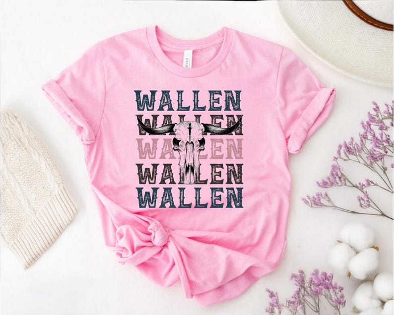 K&ouml;nnte beinhalten: Ein pinkfarbenes T-Shirt mit dem Wort "WALLEN" viermal wiederholt und einer Bullensch&auml;del-Grafik in der Mitte. Der Text ist in einer Schriftart im Western-Stil mit schwarzen, grauen und rosa Variationen. Das Shirt ist aus weichem Material.