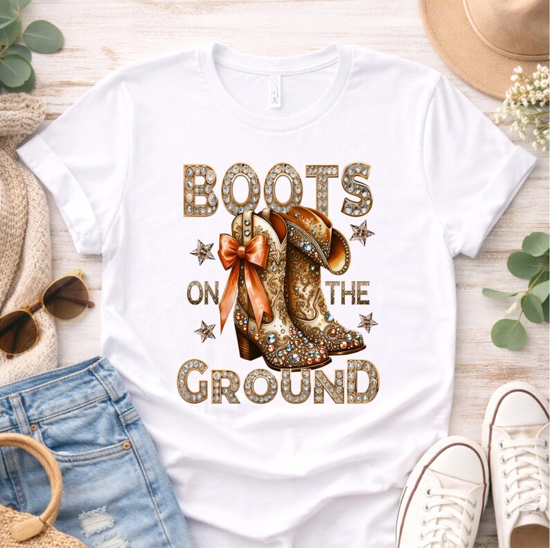 May include: White t-shirt featuring a graphic of cowboy boots with the text "BOOTS ON THE GROUND" in a decorative font. The boots are adorned with jewels and a bow. The design includes star accents.