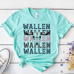 K&ouml;nnte beinhalten: Ein hellt&uuml;rkisfarbenes T-Shirt mit dem Wort "WALLEN", das viermal gestapelt ist, mit einer Bullensch&auml;del-Grafik in der Mitte. Das Wort "WALLEN" ist in Schwarz und Rosa gehalten. Ein wei&szlig;er Sneaker ist in der unteren linken Ecke zu sehen.