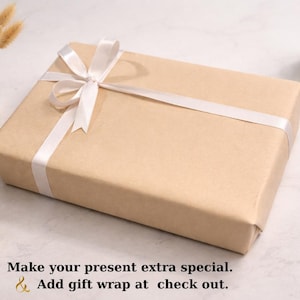 K&ouml;nnte beinhalten: Eine rechteckige Geschenkbox, verpackt in braunem Papier und mit einem wei&szlig;en Satinband gebunden. Die Box befindet sich auf einer wei&szlig;en Oberfl&auml;che, mit einer kleinen Vase mit getrockneten Blumen, einer Kerze, einem Stift und einer kleinen Schale mit Schmuck.