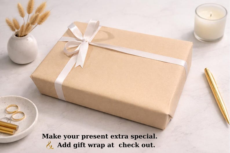 K&ouml;nnte beinhalten: Eine rechteckige Geschenkbox, verpackt in braunem Papier und mit einem wei&szlig;en Satinband gebunden. Die Box befindet sich auf einer wei&szlig;en Oberfl&auml;che, mit einer kleinen Vase mit getrockneten Blumen, einer Kerze, einem Stift und einer kleinen Schale mit Schmuck.