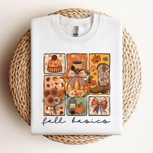 Op de afbeelding: Wit sweatshirt met een herfstthema. Het ontwerp toont een pompoen, koffiekopje, cowboylaars en andere herfst elementen. De woorden "fall basics" staan in een script lettertype onder de afbeelding.