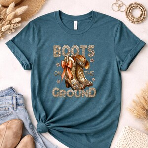 May include: Teal t-shirt featuring a graphic of cowboy boots with a bow and the text "BOOTS ON THE GROUND" in gold lettering. The boots are detailed with a floral design and rhinestones. The shirt is paired with blue jeans.