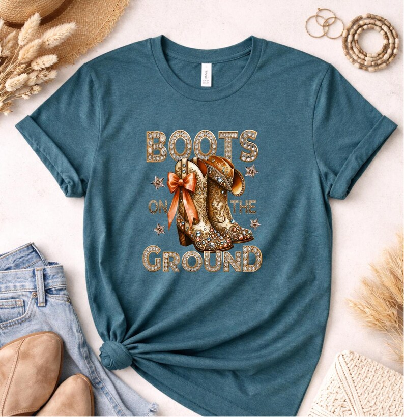 May include: Teal t-shirt featuring a graphic of cowboy boots with a bow and the text "BOOTS ON THE GROUND" in gold lettering. The boots are detailed with a floral design and rhinestones. The shirt is paired with blue jeans.