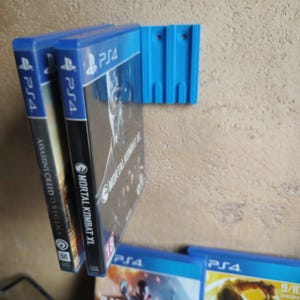 May include: A blue 3D-printed wall mount holding three PS4 game cases. The cases are labelled "Mortal Kombat XL" and "Assassin's Creed Origins". The mount is fixed to a textured wall, with more game cases visible below.