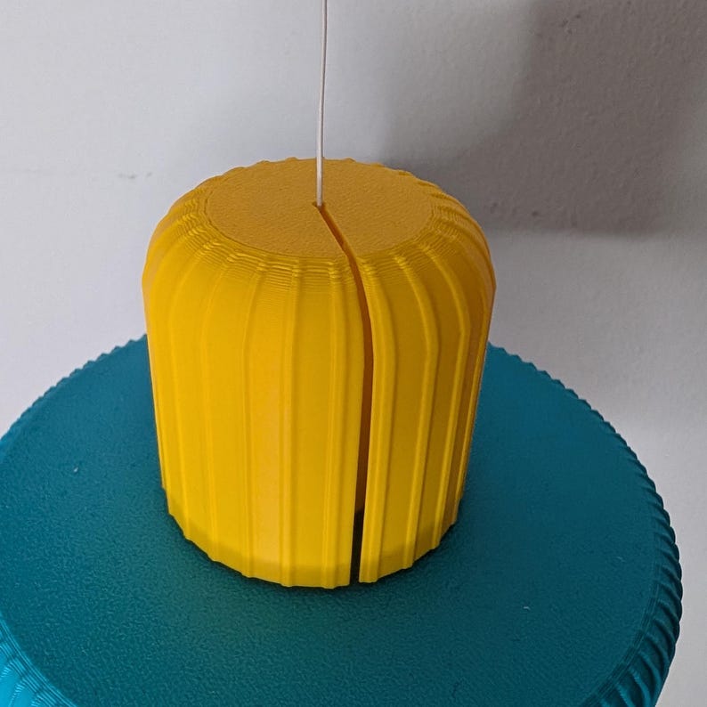 Op de afbeelding: Een heldergeel, cilindrisch object met verticale groeven en een spleet in het midden. Een dunne witte draad komt van bovenaf. Het rust op een turquoise, getextureerd oppervlak. Het ontwerp is eenvoudig en modern.