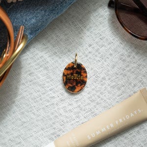 May include: An oval-shaped pendant featuring a brown and black leopard print pattern with the name "nellie" embossed in gold lettering. The pendant includes a small gold jump ring at the top for attachment, showcasing a glossy finish and distinct spots.